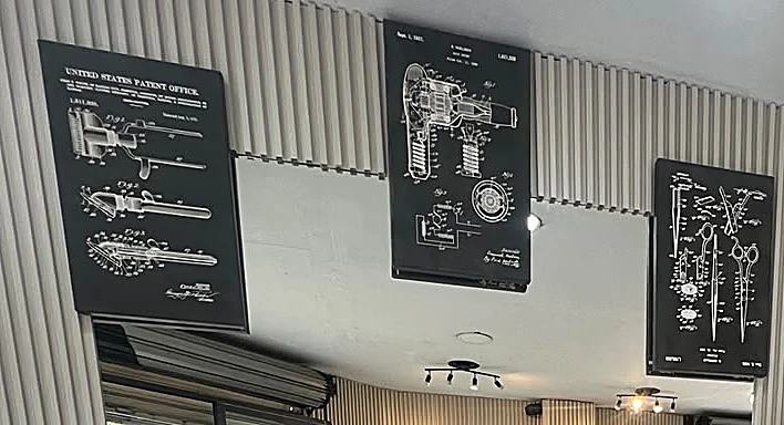 Cuadros con patentes antiguas de una máquina de cortar, una secadora y unas tijeras, sobre el muro de listones.