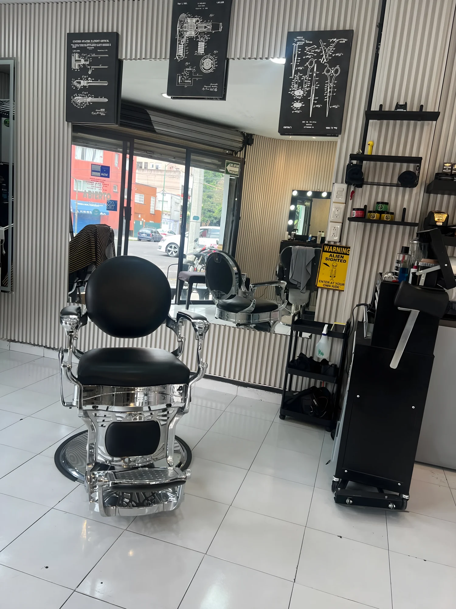 Interior de MG Barbershop en Torres Adalid: silla de barbero cromada al frente, ventana a la calle, cuadros de patentes antiguas y letrero amarillo de Alien sighted.
